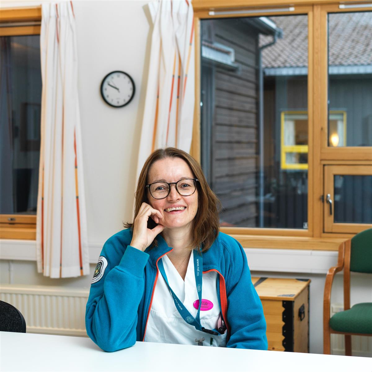 Portrett av Hilde Johansen. Bildet er tatt ved et bord inne på Prestegårdstunet sykehjem. Hilde ser i kamera og smiler, ut av vinduet bak ser man utsiden av sykehjemmet. - Klikk for stort bilde