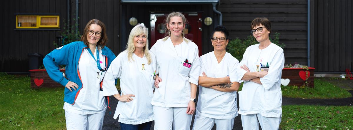 Gruppebilde av ansatte på Prestegårdstunet sykehjem. Bildet er tatt utenfor inngangsdøra til sykehjemmet, og de 5 ansatte ser i kamera og smiler. - Klikk for stort bilde