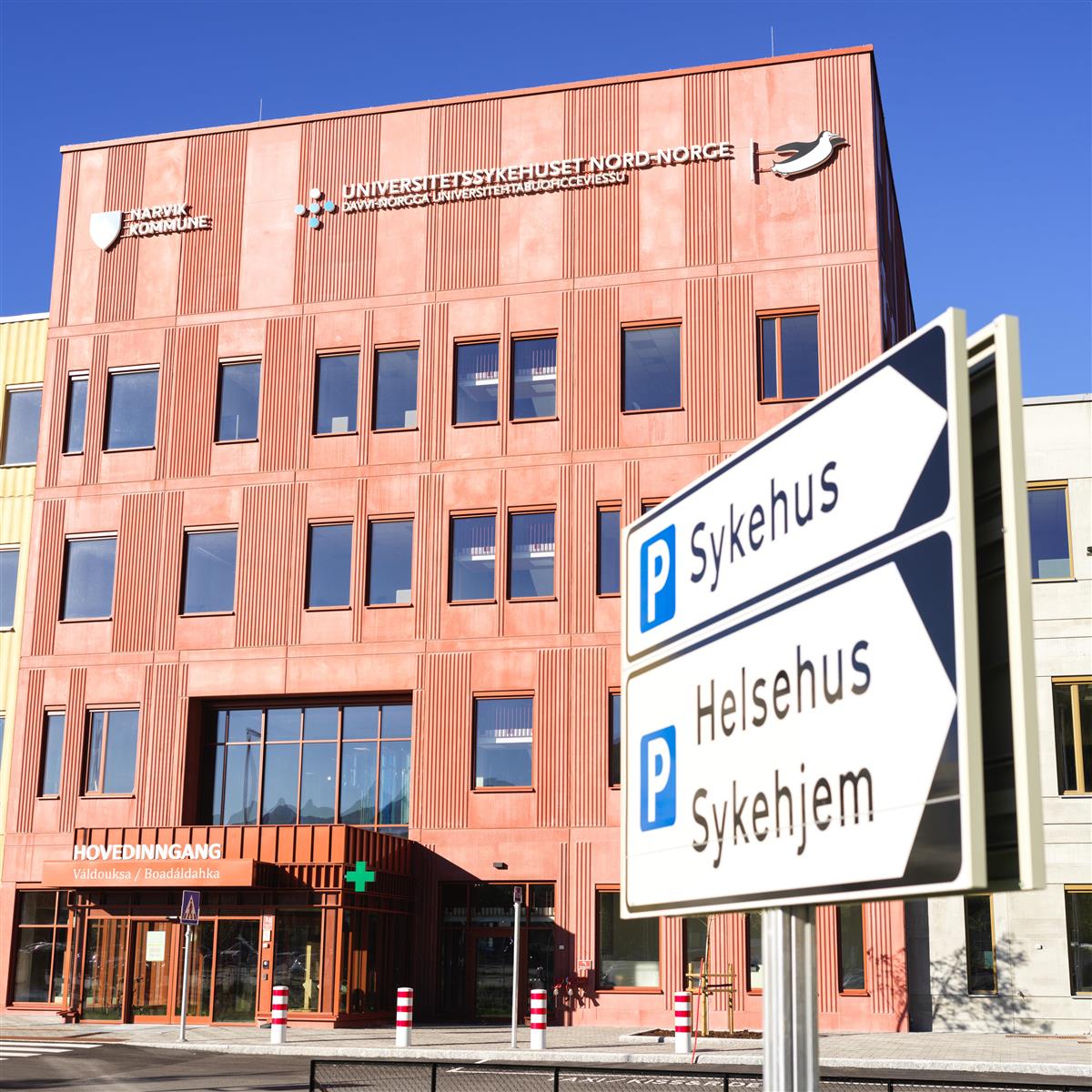 Bilde av hovedinngangen på nye UNN Narvik. I forgrunnen er det et skilt med teksten SYkehus, Helsehus og sykehjem, og peker til parkeringsplassene. - Klikk for stort bilde